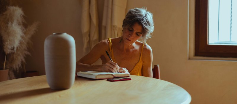 Woman writing at a table in soft light, symbolising reflection, self-awareness, and emotional healing.